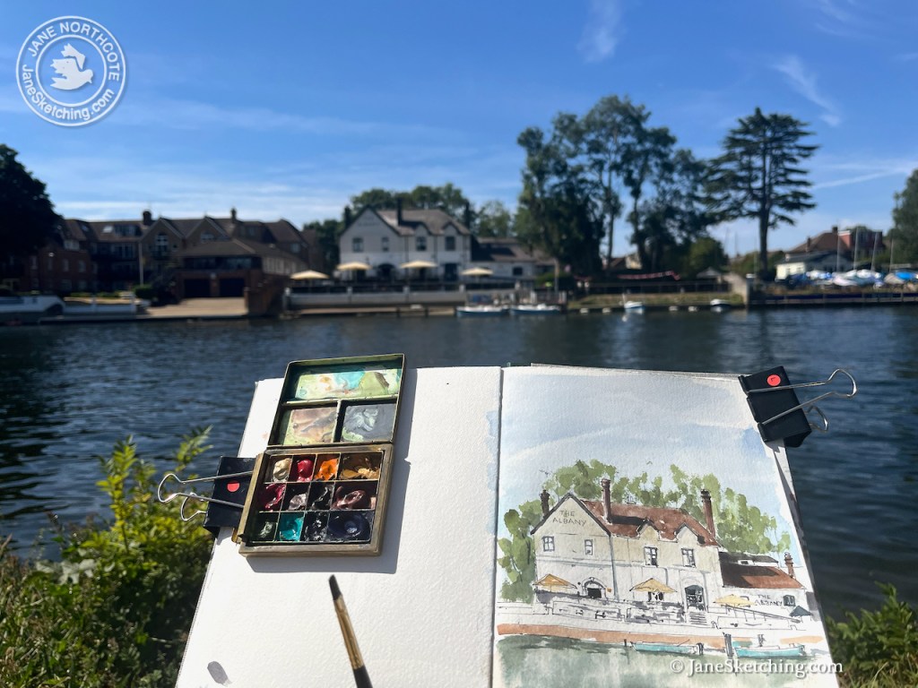A painter's watercolor palette and sketchbook displaying a painting of a riverside building titled 'The Albany,' with a scenic view of trees and boats in the background.