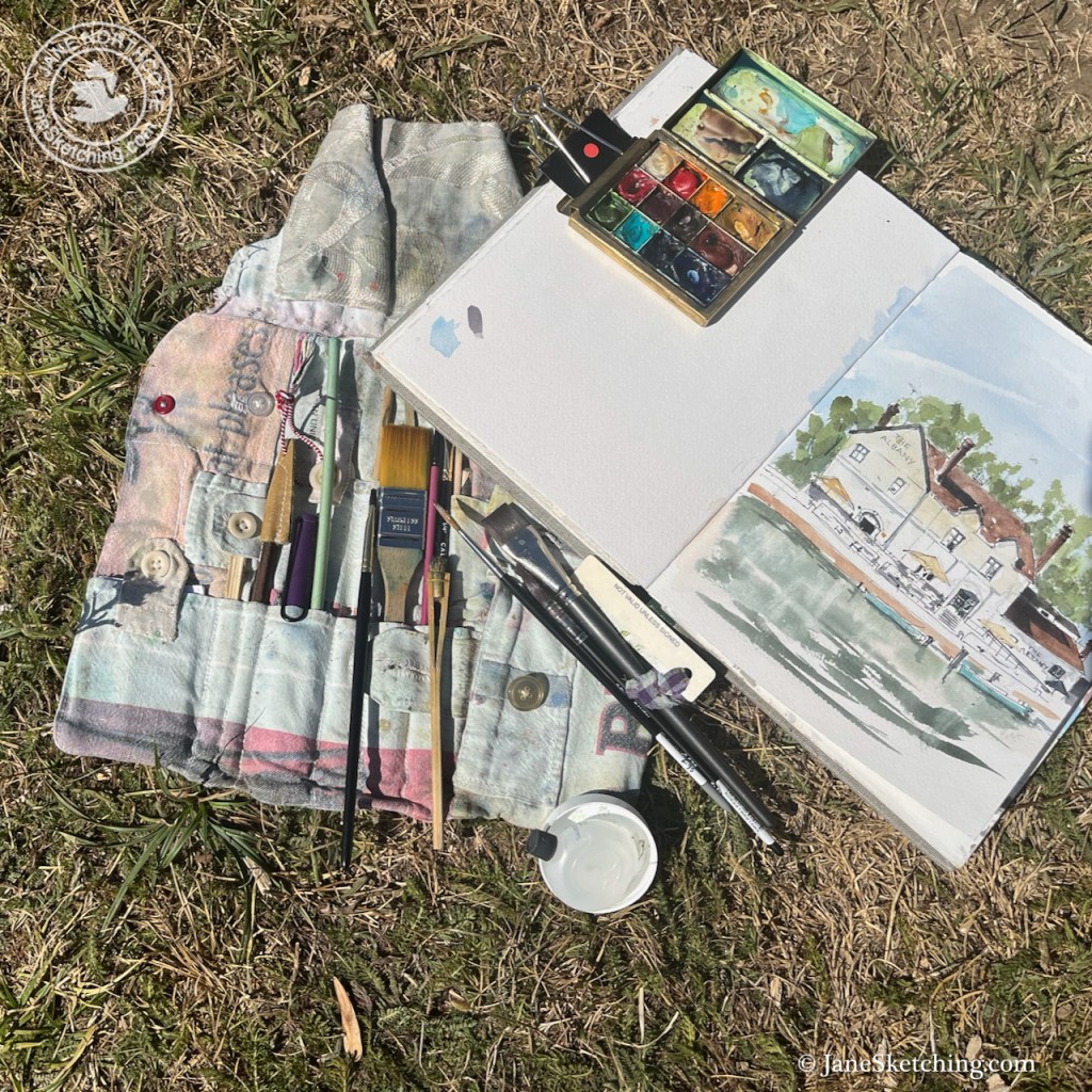 An artist's toolkit laid out on grass, featuring paintbrushes, a watercolor palette, and a sketchbook with an illustration of a house by the water.