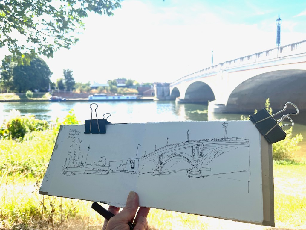 A hand holding a sketch of a bridge, with the actual bridge and river visible in the background, framed by greenery.