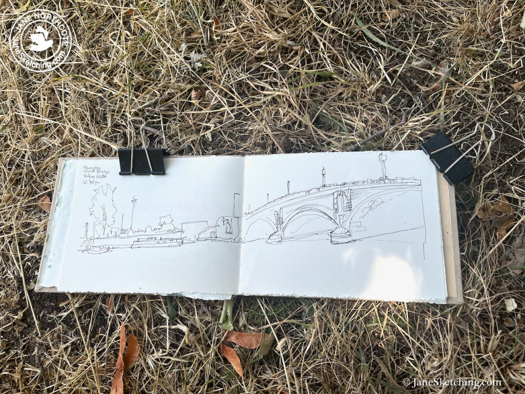 Sketchbook lying on dry grass showing a pen drawing of a bridge and surrounding scenery, secured with binder clips.